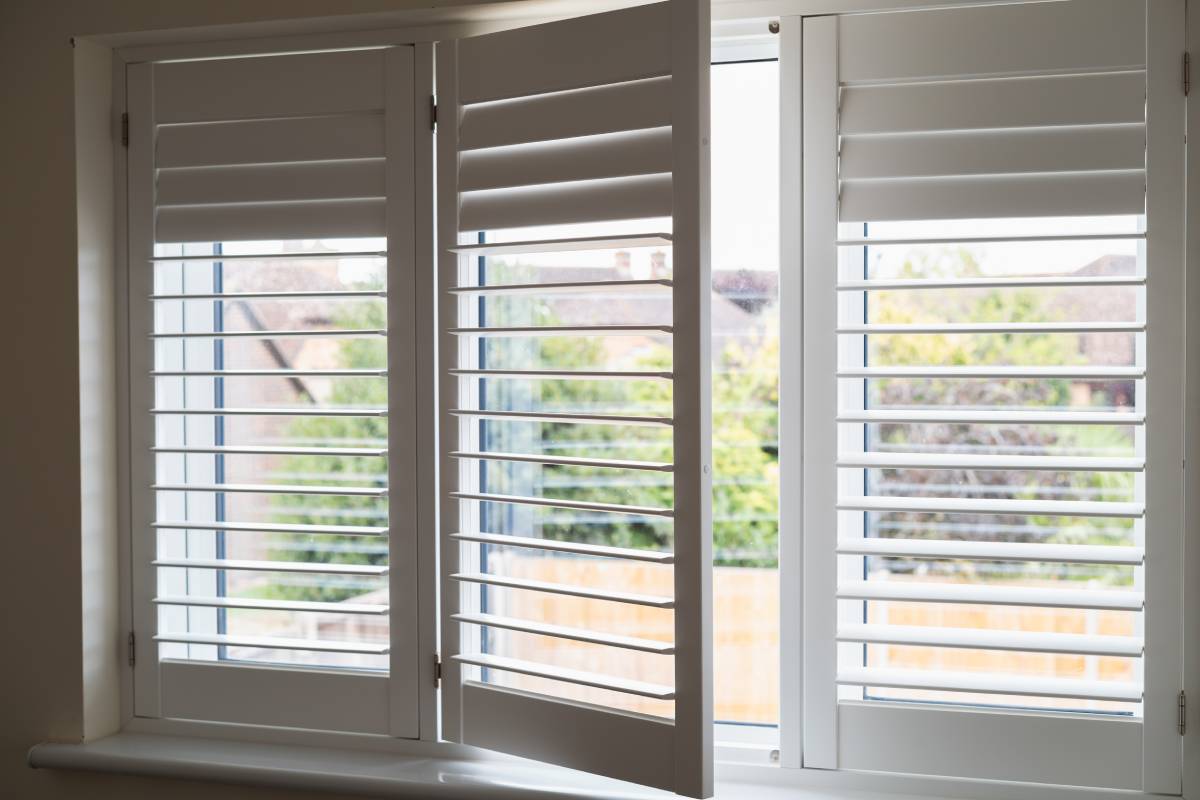 An image of white interior shutters on windows, partially closed, overlooking a fenced-in backyard near Dayton, OH