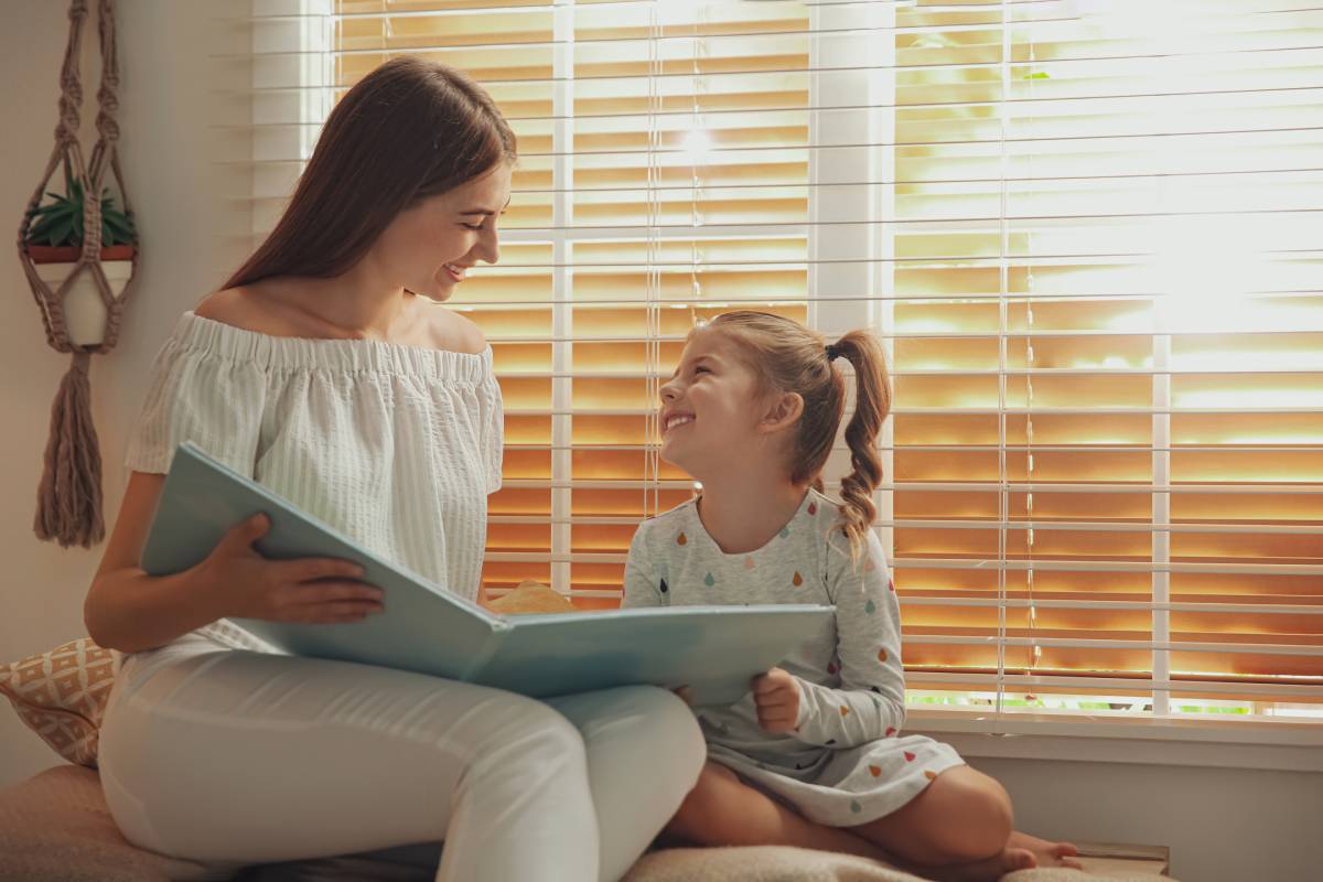 A mother and daughter sitting and reading a book together near childproof blinds at the window at Signature Shutters & Blinds near Dayton, OH