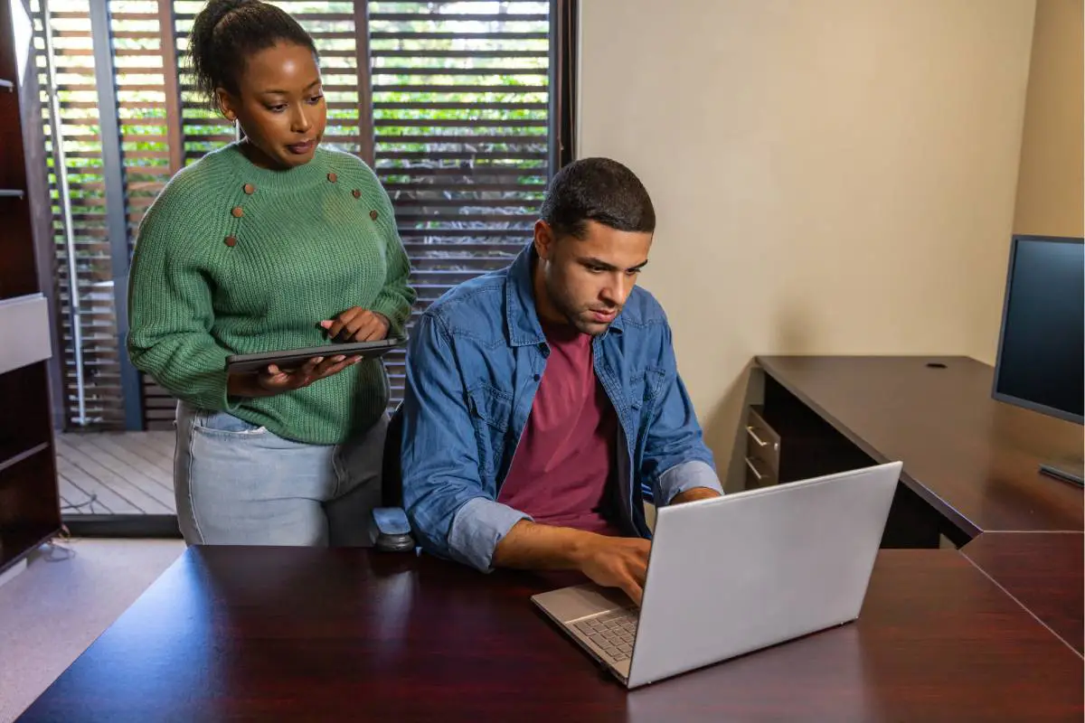 team-members-collaborating-in-a-commercial-office-space-with-interior-shutters-in-the-background-1200px | Signature Shutters and Blinds Two coworkers collaborate: a woman standing with a tablet guiding a man who is typing on a silver laptop at a dark wood desk in an office.