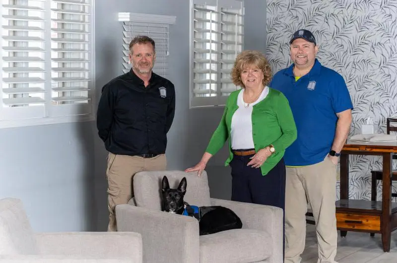 signature-shutters-about-our-company | Signature Shutters and Blinds Three adults stand in a bright living room with a small black dog sitting in a gray chair before them.